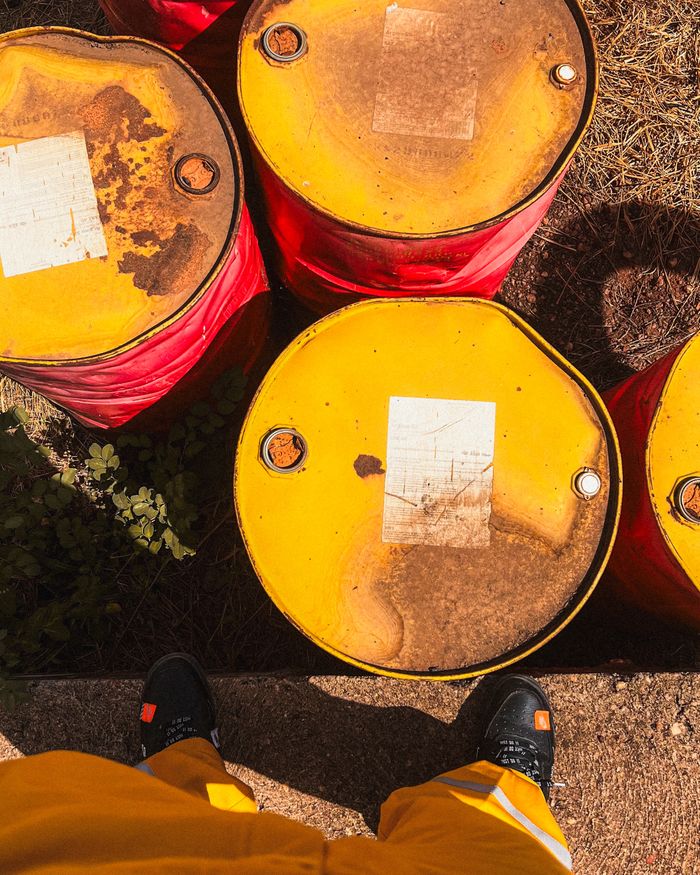 About Top view of weathered yellow oil barrels on ground, person visible wearing yellow pants.